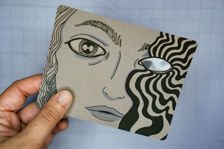 A hand holding a card with an illustration of a face with an eye that has strange radiating wiggly lines coming out of it