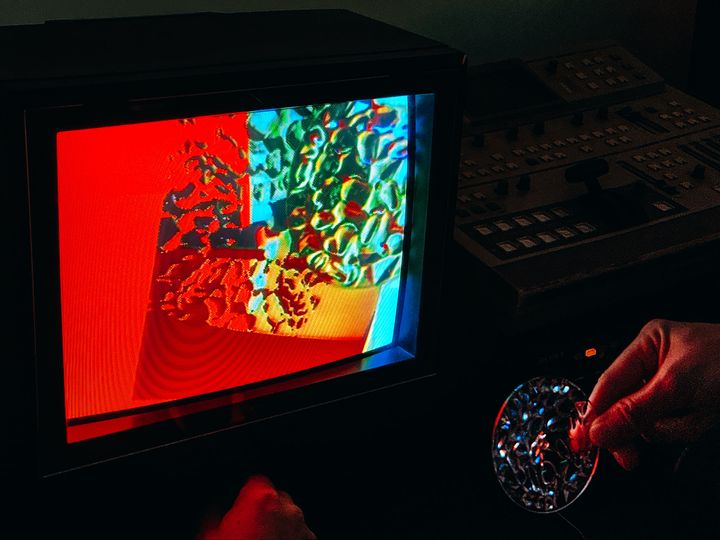 A CRT TV displaying colorful video art, a hand holding a patterned lens which is showing up on the screen